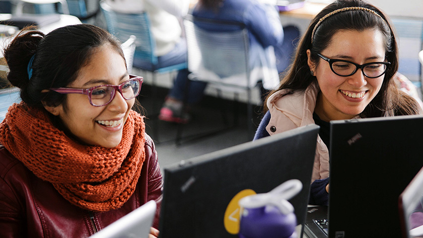 Two young women working at laptops and smiling broadly.