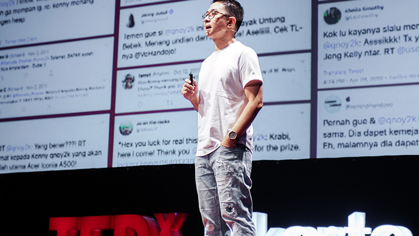 A speaker presents at TEDxJakarata with a wall of social media posts projected in the bakground